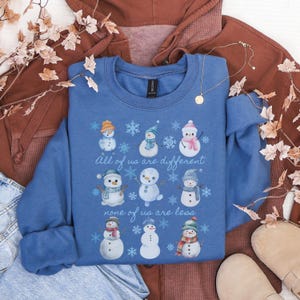 May include: A blue crewneck sweatshirt featuring a graphic of nine unique snowmen in various hats and scarves, surrounded by snowflakes. The text reads, All of us are different, none of us are less, printed in a white script font across the center.