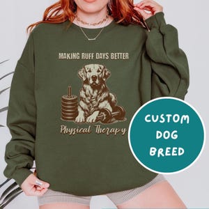 May include: Green sweatshirt with a brown illustration of a dog lifting weights. The text on the sweatshirt reads "Making Ruff Days Better" and "Physical Therapy". The text "Custom Dog Breed" is in a teal circle.