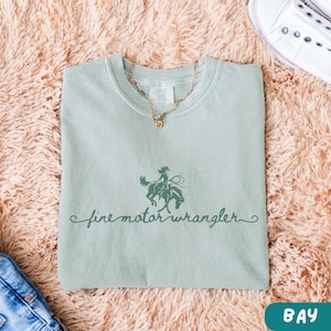 May include: A light green t-shirt with a graphic of a cowgirl riding a horse and the text "fine motor wrangler".