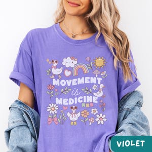 May include: Purple t-shirt with a graphic design featuring a rainbow, clouds, flowers, and ducks. The text "Movement is Medicine" is printed on the shirt.