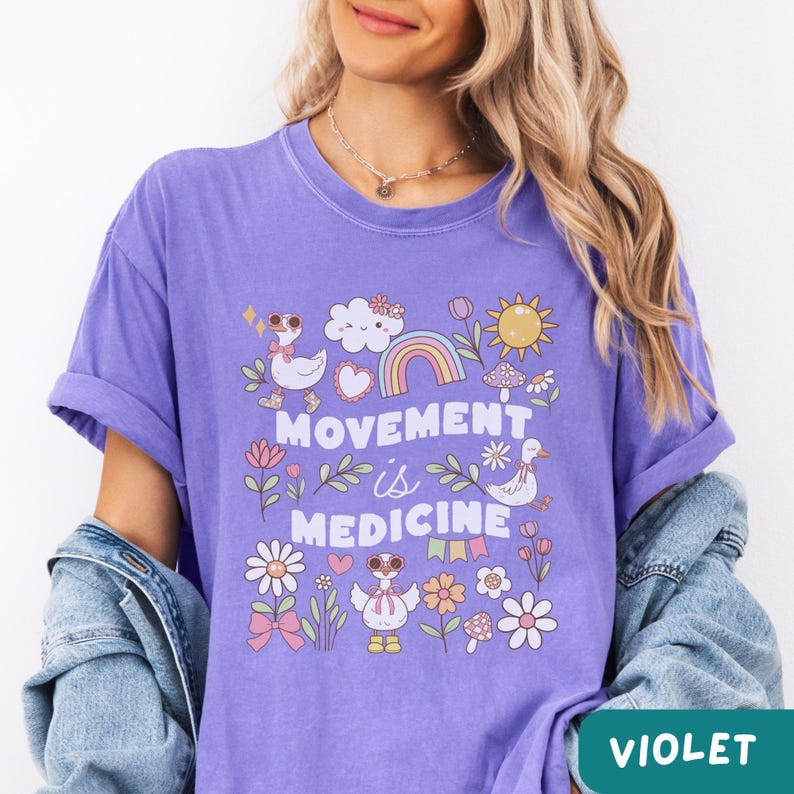 May include: Purple t-shirt with a graphic design featuring a rainbow, clouds, flowers, and ducks. The text "Movement is Medicine" is printed on the shirt.