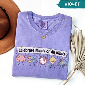 May include: A violet crewneck t-shirt featuring a graphic print with the text "Celebrate Minds of All Kinds" above illustrations of a brain, an infinity symbol, a timer, a lightbulb, and a fidget spinner, all with small smiling faces.