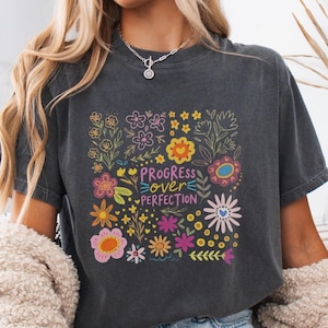 Puede incluir: Camiseta gris oscuro con un diseño floral y el texto "Progress Over Perfection" en una fuente colorida. Las flores son en tonos de rosa, amarillo, naranja y azul, creando una estética vibrante y alegre.