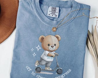 May include: A light blue Comfort Colors t-shirt featuring a cartoon teddy bear riding a scooter. The bear wears a shirt with "SPED" on it. The text "off to cheer on progress" is printed below the bear. A gold necklace is draped across the shirt.