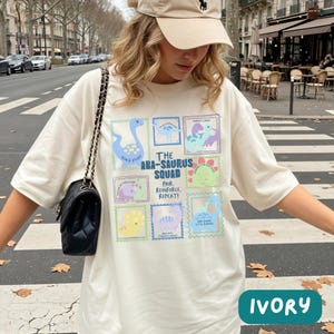 May include: An ivory t-shirt featuring a graphic print of various colorful dinosaurs in postage stamp-style frames. The text reads THE ABA-SAURUS SQUAD PAIR, REINFORCE, REPEAT! along with smaller labels under each dinosaur illustration.