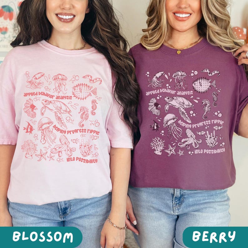 May include: Two t-shirts, one blossom pink and one berry-colored, each with a marine life graphic. The text on the shirts reads "applied behavior analysis," "helping progress ripple," and "into possibility." The shirts are paired with blue denim jeans.