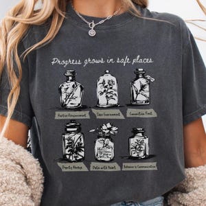 May include: Dark grey t-shirt with the text "Progress grows in safe places." The shirt features six illustrations of glass jars with botanical elements and labels: Positive Reinforcement, Safe Environment, Connection First, Dignity Always, Data with Heart, and Behavior is Communication.