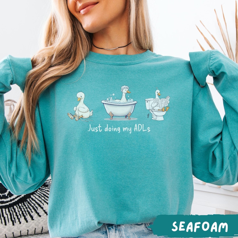 Op de afbeelding: Zeegroen sweatshirt met een afbeelding van drie cartoon eenden in verschillende badkameractiviteiten. De tekst "Just doing my ADLs" staat onder de eenden. De sweatshirt heeft lange mouwen en een ronde hals.