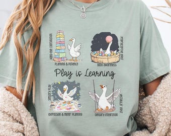 May include: Sage green t-shirt with a graphic featuring a goose in various playful scenarios, including building blocks and a swing. The text "Play is Learning" is prominently displayed, along with related terms like "Hand-eye coordination" and "Sensory Play".