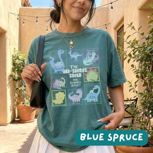 May include: A dark teal short-sleeve t-shirt featuring a graphic print of various dinosaurs in rectangular frames. The text reads THE ABA-SAURUS SQUAD PAIR, REINFORCE, REPEAT! The shirt is worn with a black shoulder bag and a gold layered necklace.