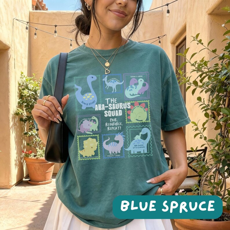May include: A dark teal short-sleeve t-shirt featuring a graphic print of various dinosaurs in rectangular frames. The text reads THE ABA-SAURUS SQUAD PAIR, REINFORCE, REPEAT! The shirt is worn with a black shoulder bag and a gold layered necklace.
