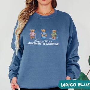 May include: Indigo blue sweatshirt featuring three cartoon bears in active poses. The bears are wearing different outfits and are engaged in activities like using a cane, exercising on a ball, and doing yoga. The text "Bear with me, Movement is Medicine" is printed on the front.