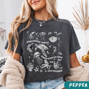 May include: Dark gray t-shirt featuring a marine life graphic with the phrases "applied behavior analysis", "helping progress ripple", and "into possibility". The design includes illustrations of jellyfish, a sea turtle, seahorses, and other sea creatures.