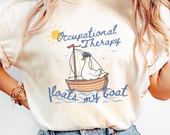 Może przedstawiać: Kremowa koszulka z napisem "Occupational Therapy floats my boat". Projekt przedstawia kreskówkową kaczkę w żaglówce, żółte słońce i niebieskie fale. Koszulka jest codziennym elementem garderoby.