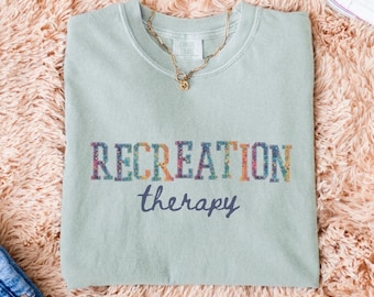 May include: A light green t-shirt with the words "RECREATION" and "therapy" printed on the front. The word "RECREATION" is in colorful floral letters. A gold necklace with a pendant is on the shirt.