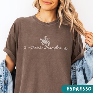 K&ouml;nnte beinhalten: Ein braunes T-Shirt mit einem Aufdruck einer Cowgirl, die ein Pferd reitet, und dem Text "crisis wrangler" in einer kursivschrift.