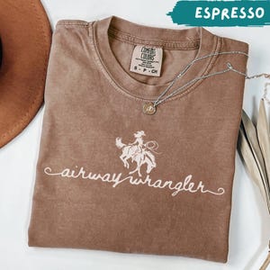 May include: A brown short-sleeve t-shirt with a crew neckline and a white graphic print of a person riding a bucking horse. The text "airway wrangler" is printed below the graphic. A silver-toned necklace with a small round pendant rests on the shirt.