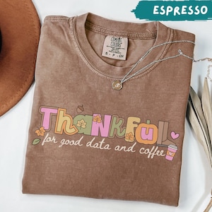 May include: A brown short-sleeve t-shirt with a crew neckline featuring the text Thankful for good data and coffee in colorful block letters. The design includes small leaf icons, an acorn, a boot, and a coffee cup. A silver chain necklace rests on the shirt.