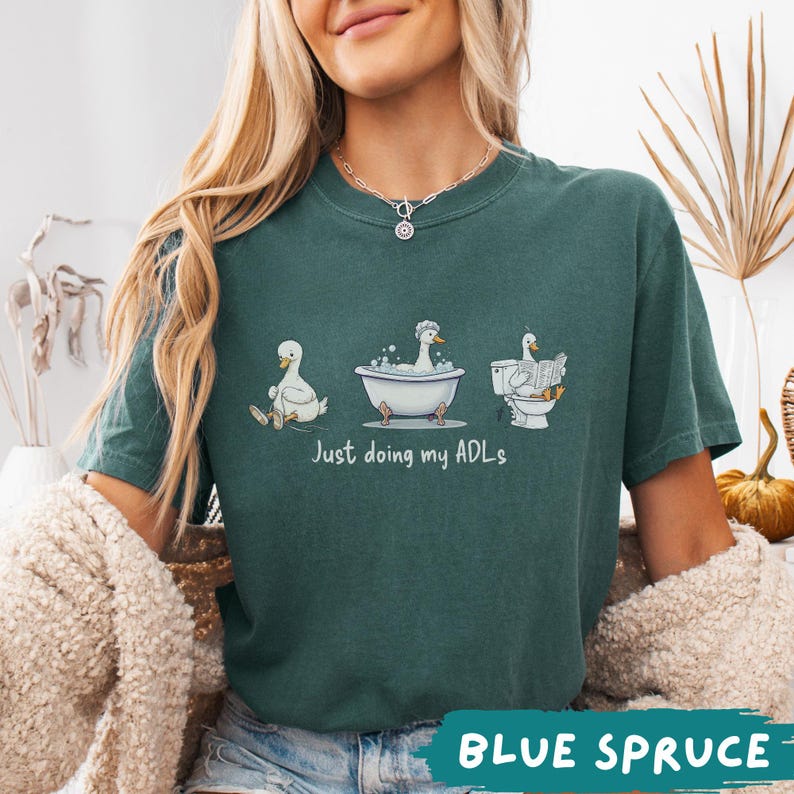 Op de afbeelding: Een tealkleurig T-shirt met een grappig ontwerp van cartoon eenden die dagelijkse activiteiten uitvoeren. De tekst "Just doing my ADLs" staat eronder. De kleur van het shirt is "BLUE SPRUCE".