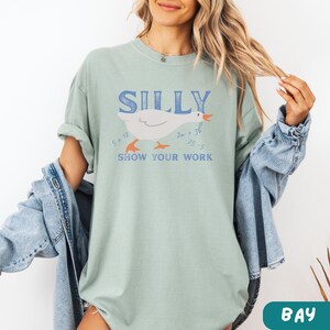 May include: A light green t-shirt with the word "SILLY" in blue block letters above a cartoon goose illustration. The goose has orange feet and beak, with math equations around it. The text "SHOW YOUR WORK" is below the goose.