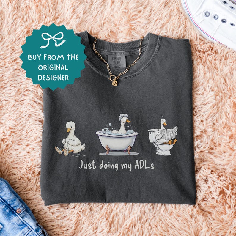Op de afbeelding: Donkergrijze t-shirt met een grappige illustratie van eenden die dagelijkse taken uitvoeren: zitten, baden en lezen. De tekst "Just doing my ADLs" staat eronder. Een gouden ketting ligt op het shirt.
