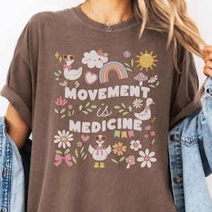 May include: A brown t-shirt with a graphic design featuring a rainbow, clouds, a sun, mushrooms, flowers, and geese. The text "Movement is Medicine" is printed on the shirt.