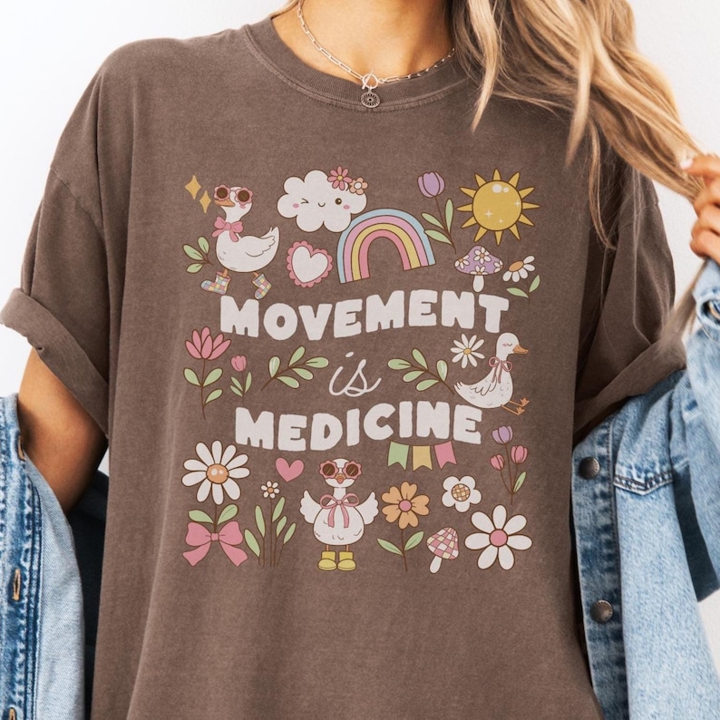 May include: A brown t-shirt with a graphic design featuring a rainbow, clouds, a sun, mushrooms, flowers, and geese. The text "Movement is Medicine" is printed on the shirt.