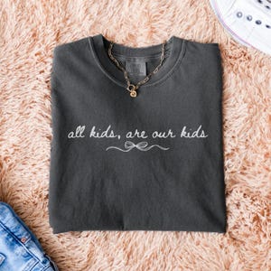 May include: Dark gray Comfort Colors t-shirt with the text "all kids, are our kids" in white script. A gold chain necklace with a pendant rests on the shirt. The shirt is folded on a pink fluffy surface.