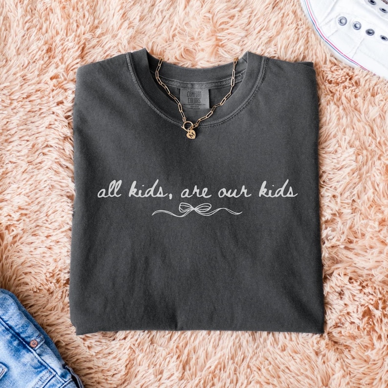 May include: Dark gray Comfort Colors t-shirt with the text "all kids, are our kids" in white script. A gold chain necklace with a pendant rests on the shirt. The shirt is folded on a pink fluffy surface.