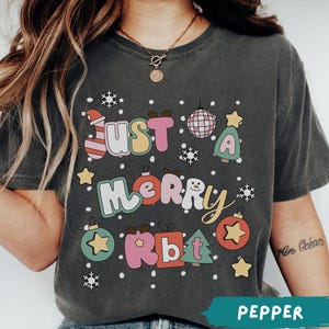 May include: A charcoal gray t-shirt featuring the text JUST A MERRY Rbt in colorful, bubble-style letters. The design includes graphic elements such as a disco ball, Christmas trees, ornaments, stars, and snowflakes scattered across the front.