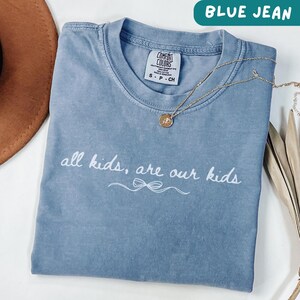 May include: Blue jean colored t-shirt with the text "all kids, are our kids" in white script. A gold necklace with a pendant is draped across the shirt. The shirt is a Comfort Colors brand, size S-P-CH.