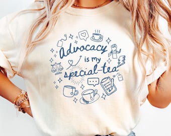 May include: A cream-coloured t-shirt featuring a blue graphic that says "Advocacy is my special-tea." The design includes teacups, hearts, scales, and other icons. The shirt is made of a soft material.