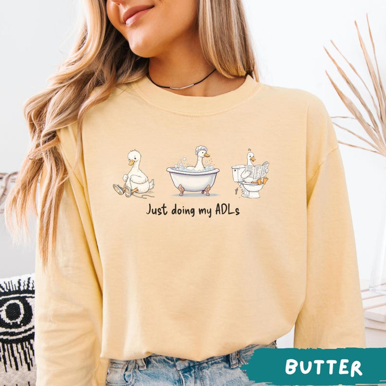 Op de afbeelding: Boterkleurige sweatshirt met een grillige afbeelding van drie eenden die dagelijkse activiteiten uitvoeren: zitten, baden en lezen. De tekst "Just doing my ADLs" staat onder de eenden. De sweatshirt heeft een ronde hals.