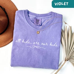 May include: A violet Comfort Colors t-shirt with the text "all kids, are our kids" in white script. A gold necklace with a pendant rests on the shirt. A brown hat and dried palm leaf are in the background.