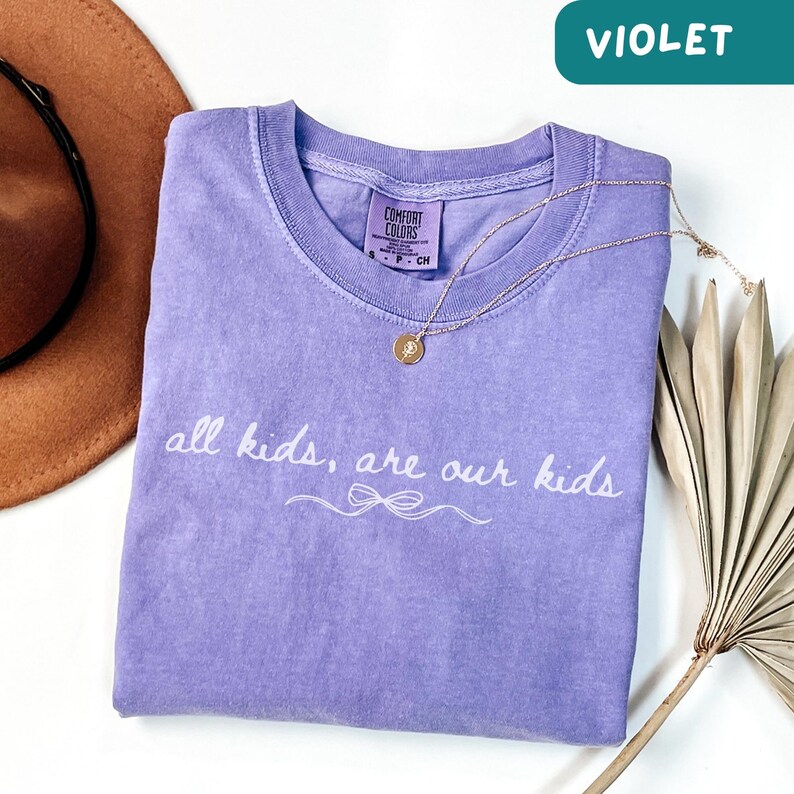 May include: A violet Comfort Colors t-shirt with the text "all kids, are our kids" in white script. A gold necklace with a pendant rests on the shirt. A brown hat and dried palm leaf are in the background.