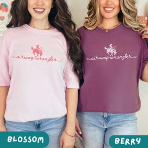 Pu&ograve; includere: Due donne indossano magliette con il testo "airway wrangler" in rosa e viola. Il testo &egrave; in un font corsivo e presenta una grafica di una cowgirl a cavallo. La donna a sinistra indossa una maglietta rosa e la donna a destra indossa una maglietta viola. Il testo "blossom" &egrave; nell'angolo in basso a sinistra e il testo "berry" &egrave; nell'angolo in basso a destra.