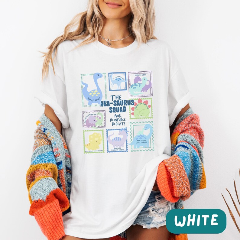 May include: A white short-sleeve t-shirt featuring a graphic print of various colorful dinosaurs arranged in rectangular frames. The text reads The ABA-SAURUS SQUAD, PAIR, REINFORCE, REPEAT! along with smaller phrases like SLOW & STEADY.