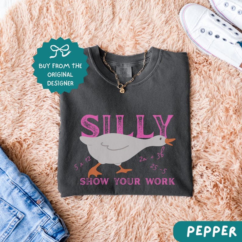 May include: Dark gray t-shirt with the word "SILLY" in pink, a cartoon goose, and the phrase "SHOW YOUR WORK" in pink. The shirt also features math equations. A gold necklace and white sneakers are visible.