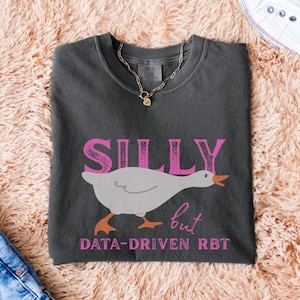 May include: Dark grey t-shirt featuring the word "SILLY" in pink block letters above a cartoon goose illustration. Below the goose is the text "but DATA-DRIVEN RBT" in pink. A gold chain necklace is draped over the shirt.