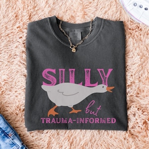 May include: Dark gray t-shirt with the words "SILLY but TRAUMA-INFORMED" in pink. A cartoon goose with orange feet and beak is featured below the word "SILLY". A gold chain necklace is draped on the shirt.