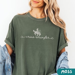 K&ouml;nnte beinhalten: Ein gr&uuml;nes T-Shirt mit einer wei&szlig;en Grafik einer Frau, die ein Pferd reitet, und dem Text "crisis wrangler".