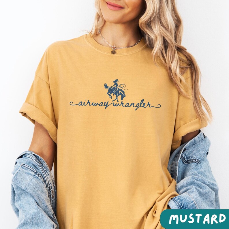 Pu&ograve; includere: Una maglietta gialla senape con una grafica blu di una cowgirl a cavallo. Il testo "airway wrangler" &egrave; stampato in blu sotto la grafica.