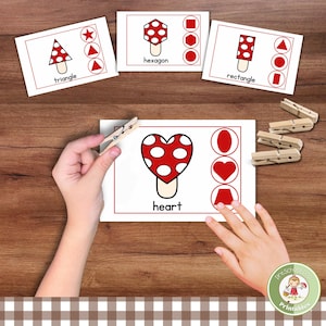 May include: Educational cards with mushroom shapes and matching shapes. The cards feature a red and white mushroom design with the shape names: triangle, hexagon, rectangle, and heart. A child's hand holds a clothespin to match the shapes.