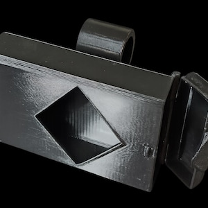 May include: A black, rectangular box with a diamond-shaped cutout and a hinged lid. A cylindrical loop is attached to the top. The lid is open, revealing the interior. The object is made of a textured material.
