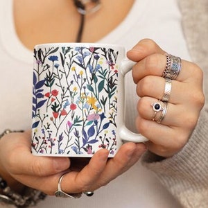 May include: A white ceramic mug with a colorful floral pattern. The mug has a handle and is being held by a person.