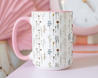 Può includere: Una tazza in ceramica bianca con manico e bordo rosa. La tazza è ricoperta da un motivo di delicati fiori di campo colorati. La tazza è appoggiata su una superficie rosa.
