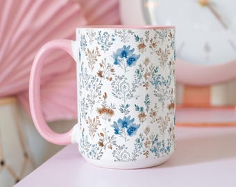 May include: A white ceramic mug with a pink handle and a floral pattern in shades of blue and brown. The mug is filled with a white liquid.