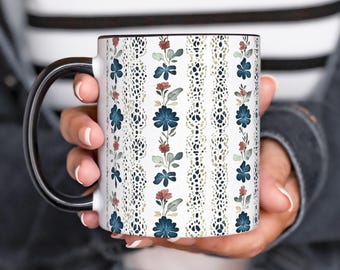 May include: A white ceramic mug with a black handle. The mug features a repeating pattern of blue and red flowers with green leaves on a white background. The pattern is framed by a thin black border.