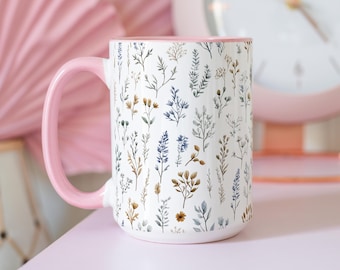 Può includere: Una tazza in ceramica bianca con un bordo e un manico rosa. La tazza presenta un motivo floreale con fiori e foglie blu e marroni.
