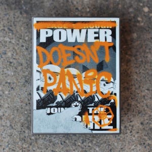 Könnte beinhalten: Ein weißer Aufkleber mit orangefarbenem Text, der "Power Doesn't Panic" lautet, mit einem Schwarzweißbild von Stormtroopern aus Star Wars. Der Aufkleber befindet sich auf einer grauen Betonfläche.